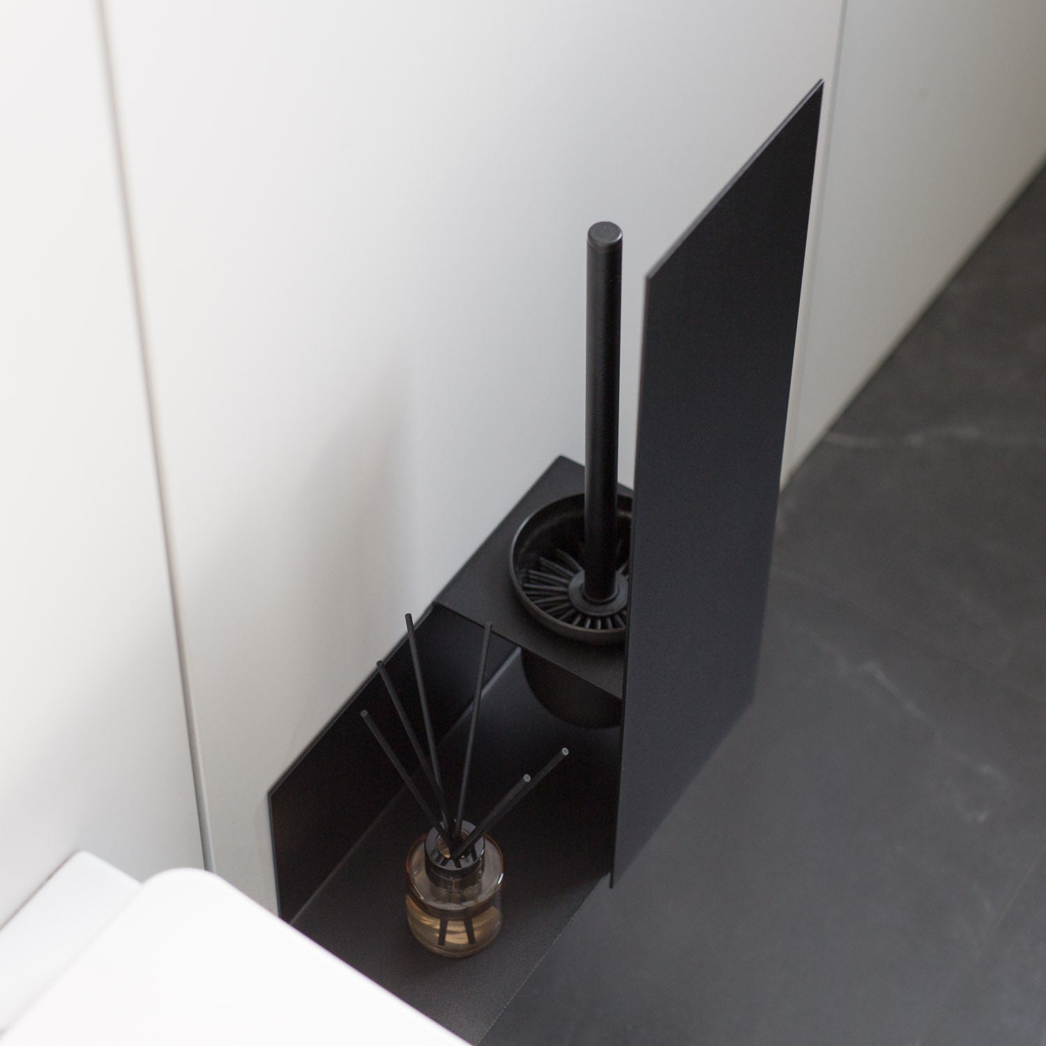 Black wall-mounted toilet brush holder with a diffuser against mounted to a white wall.