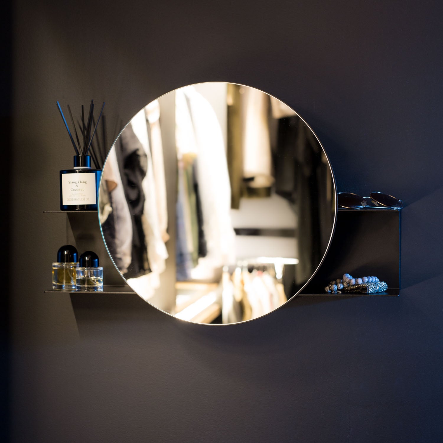 Round black wall mirror with shelf on a dark wall with decorative items around it.