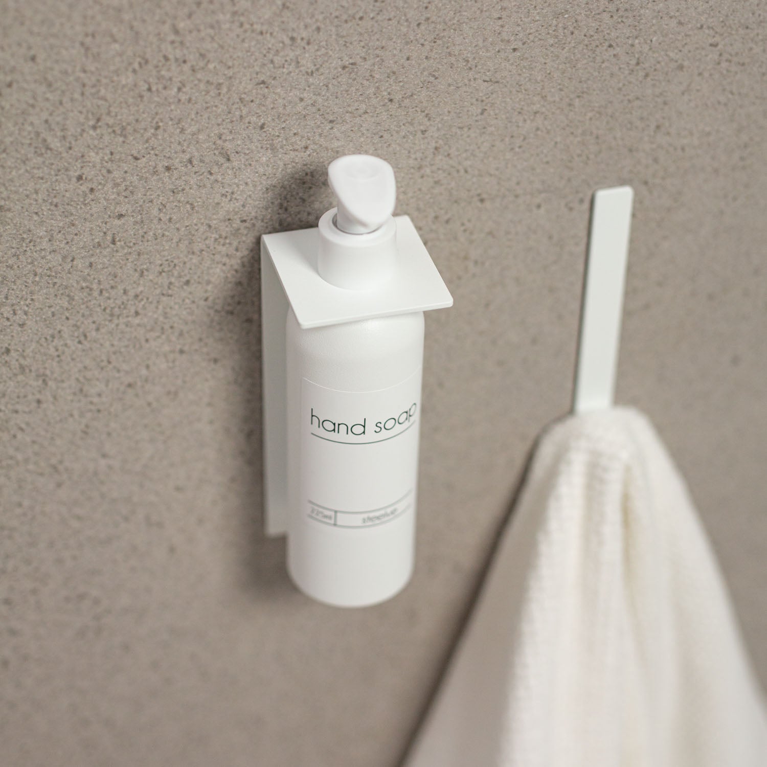 White hand soap dispenser mounted on a beige wall with a white towel hanging nearby.