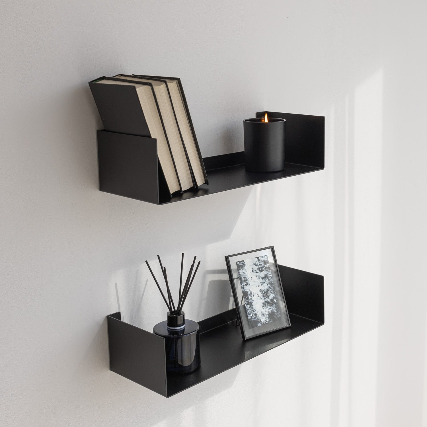 Black modern wall shelves with books, a candle, and decorative items against a white wall.