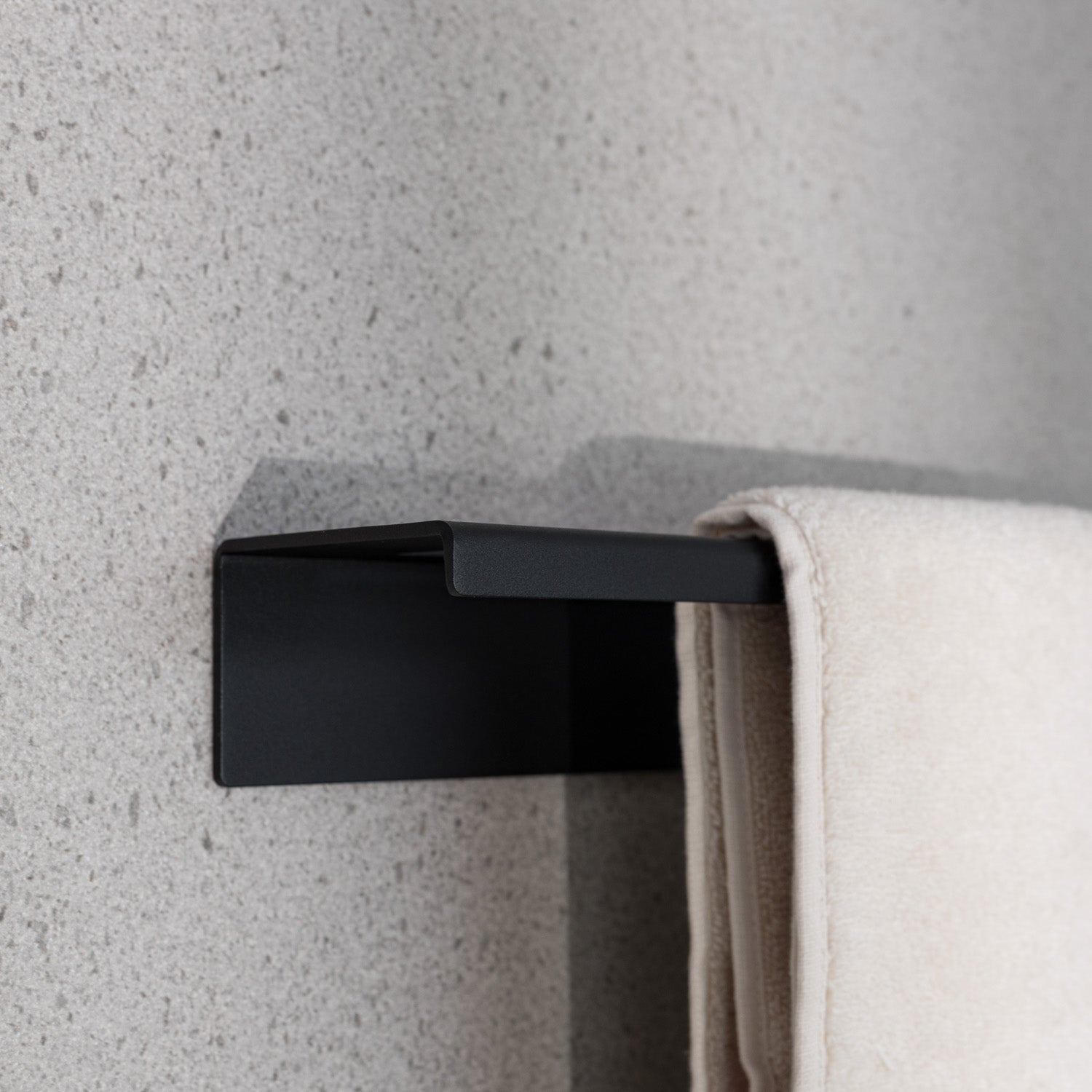 no-drill Black towel hook on a textured gray wall with a beige towel draped over it.