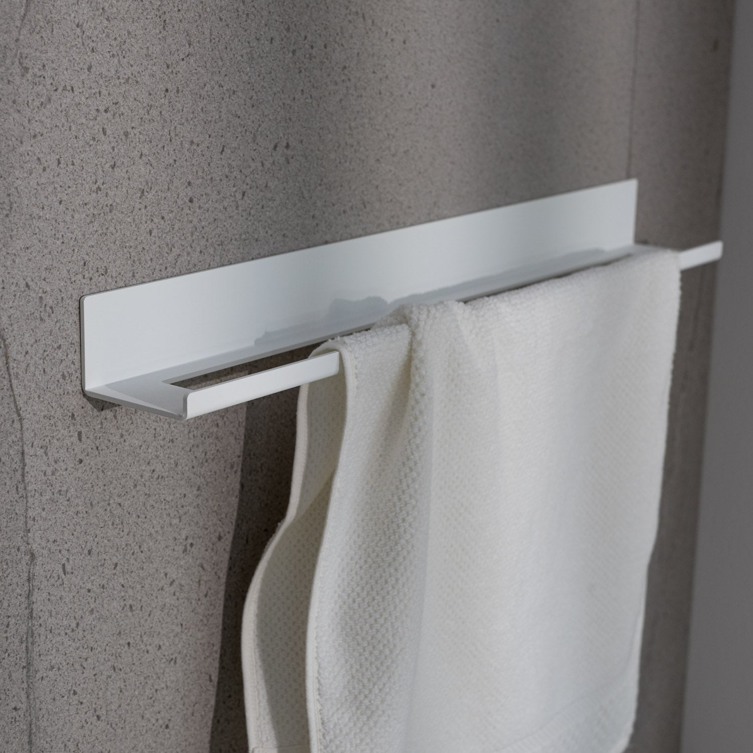Small White towel bar with towel mounted against a gray concrete wall.